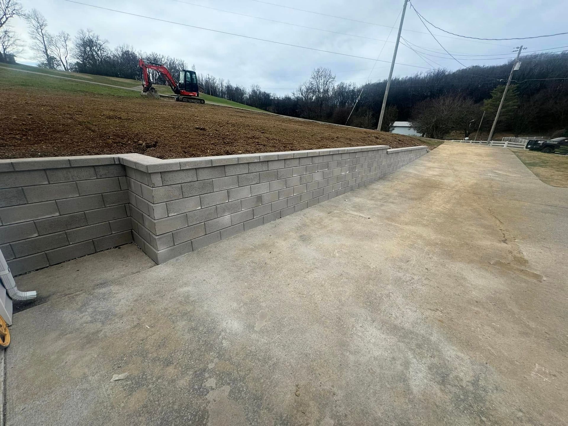Concrete retaining wall next to a paved area, with an excavator on a hillside in the background under a cloudy sky.