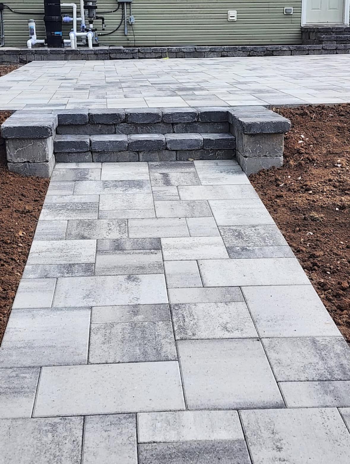A stone pathway leads to steps and a patio area, constructed of grey pavers. The setting is outdoors, with some exposed soil.