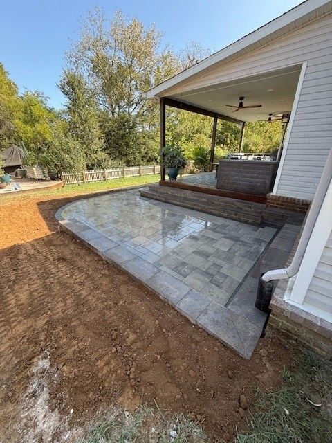 Paver patio beside a house with a covered porch, and brown soil surrounds the patio.