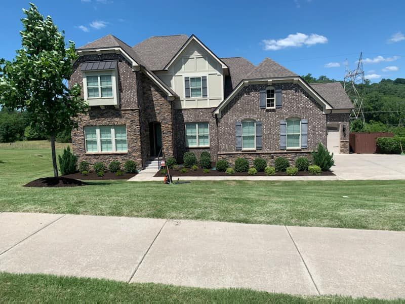 Two-story brick home with tan accents, shutters, and a well-manicured lawn under a blue sky.