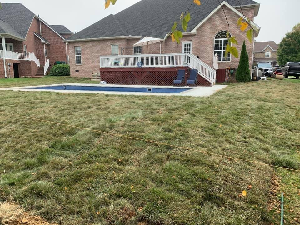 Backyard view with a pool and deck, surrounded by patchy grass and two-story brick houses under a gray sky.