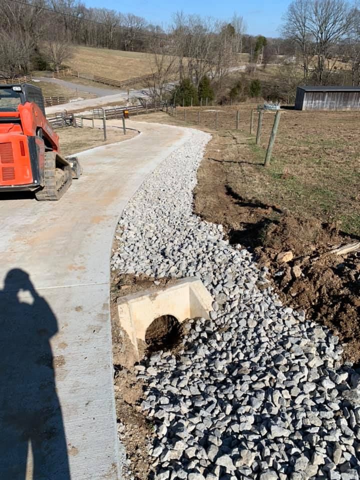 A gravel drainage system being constructed beside a concrete driveway, with a small culvert.