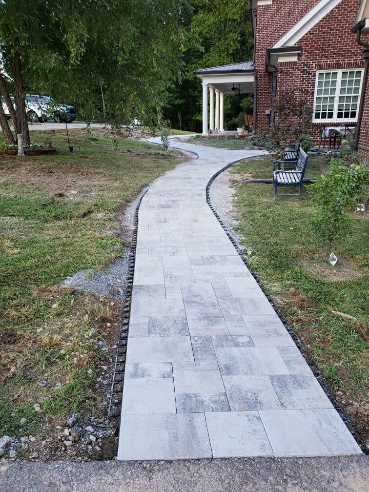 Stone walkway curves toward a brick house with white columns. A bench sits nearby on the grass.