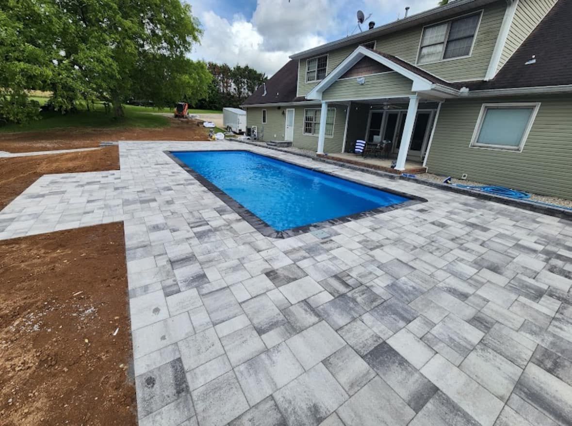 A rectangular pool with blue water is surrounded by gray pavers. The pool is beside a two-story green house on a sunny day.