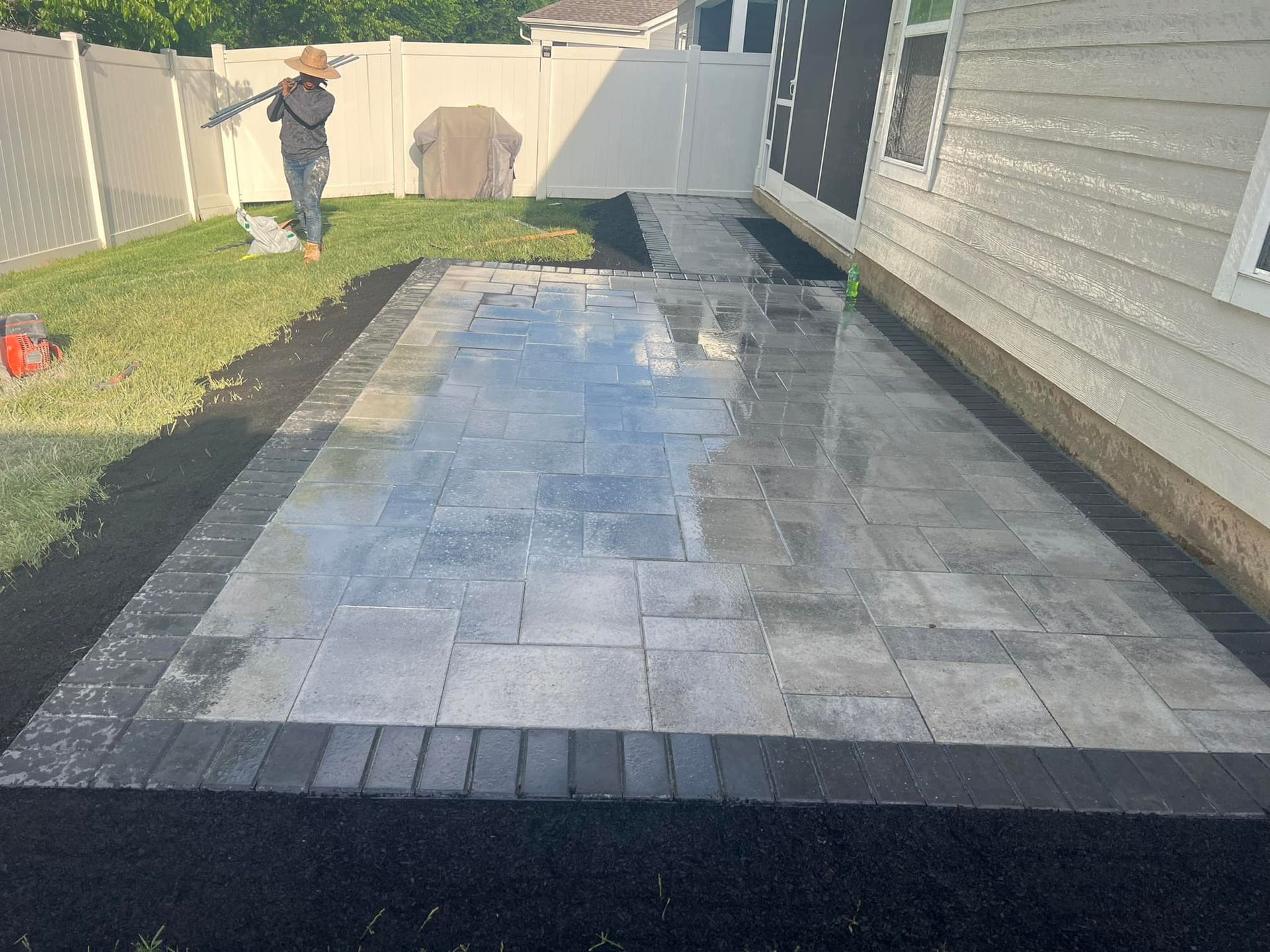 Newly installed gray paver patio with a dark border. A person works in the background next to a white fence.