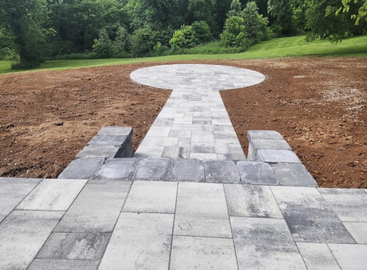 Gray stone patio and pathway leading to a circular platform on a grassy hill, surrounded by dirt and trees.