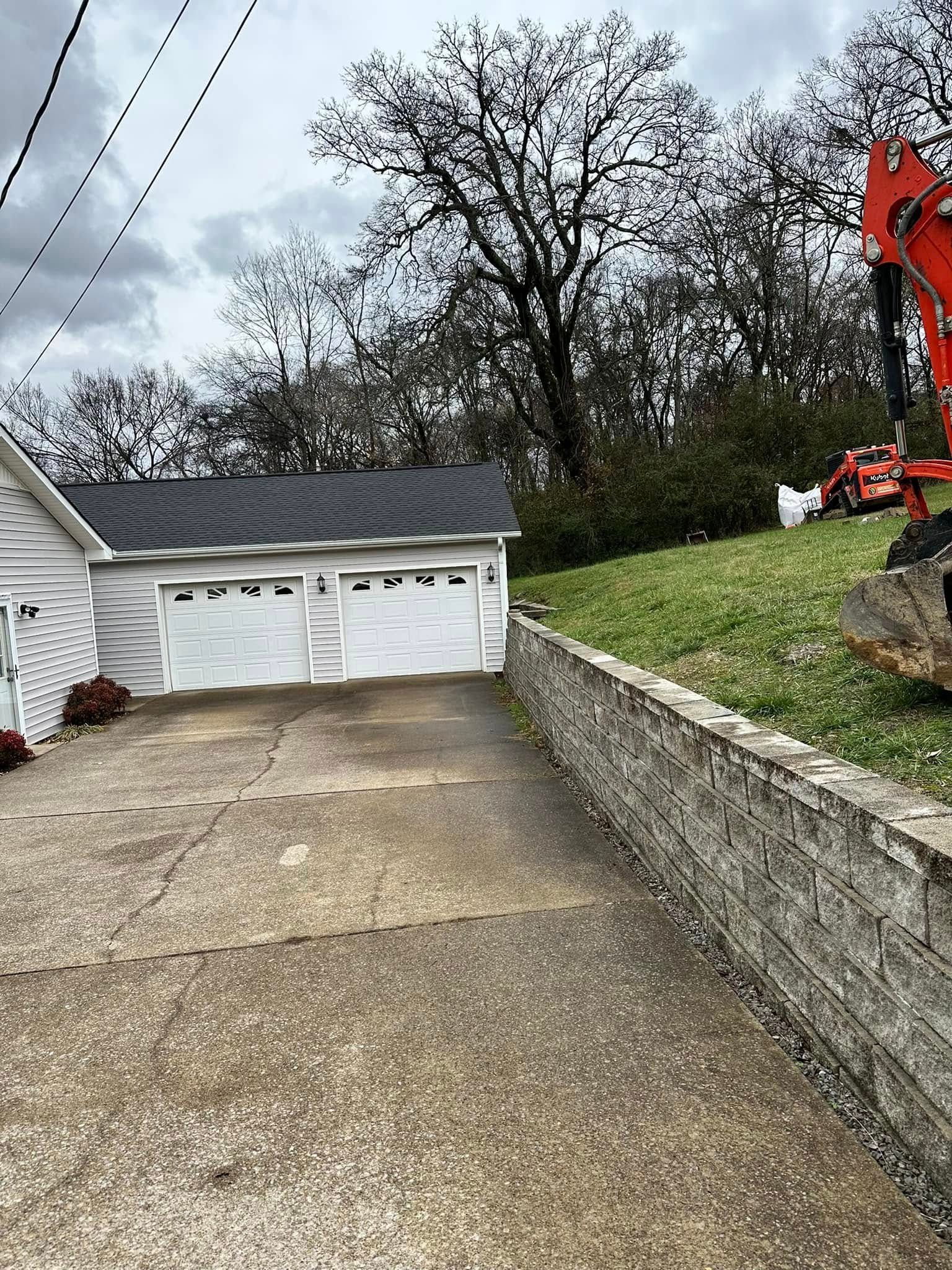 A concrete driveway slopes downward toward a two-car garage. A retaining wall and hillside are to the right.