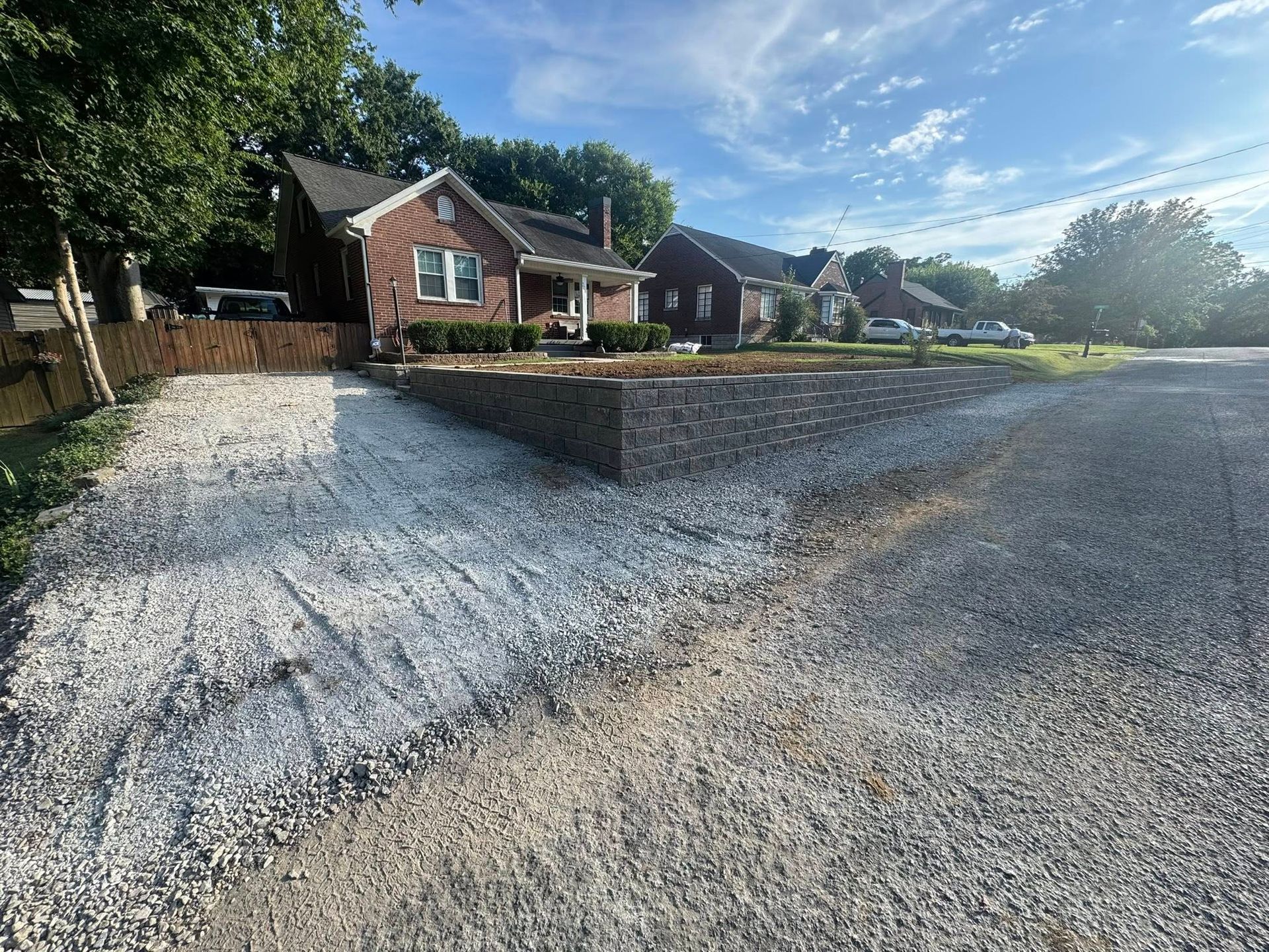 Gravel driveway in front of brick houses.  Sunlit, with a gabion retaining wall and trees lining the horizon.