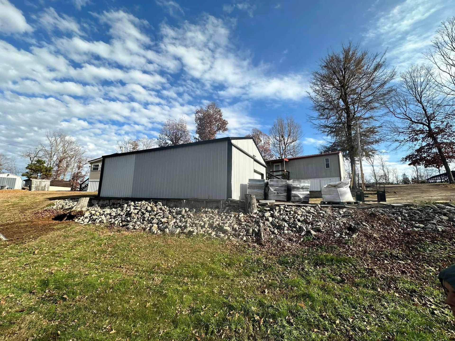 A metal-sided building on a rock foundation, with construction materials stacked nearby under a cloudy sky.