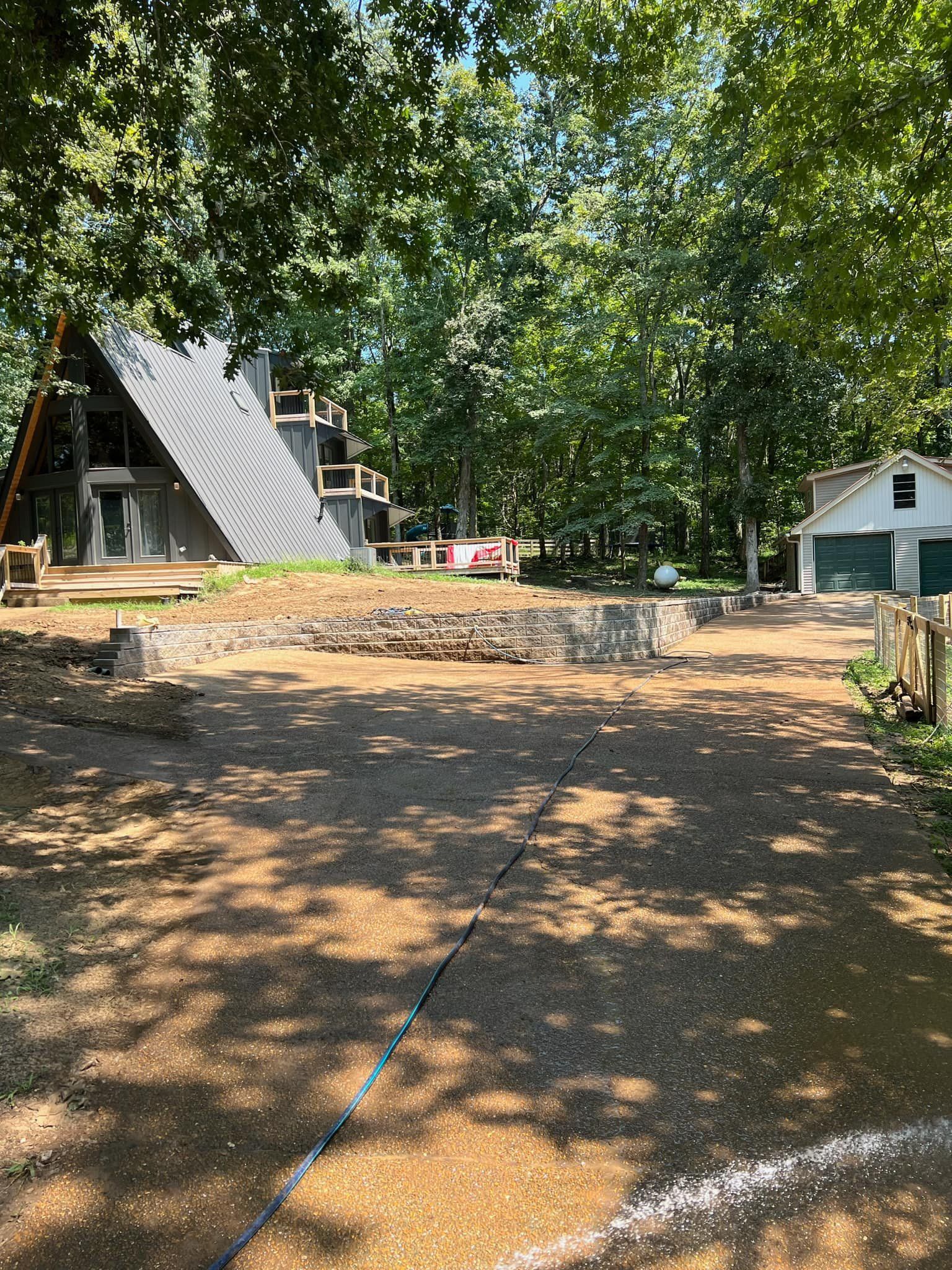 A-frame house nestled in a forest with a gravel driveway and detached garage. Sunny day with shadows.