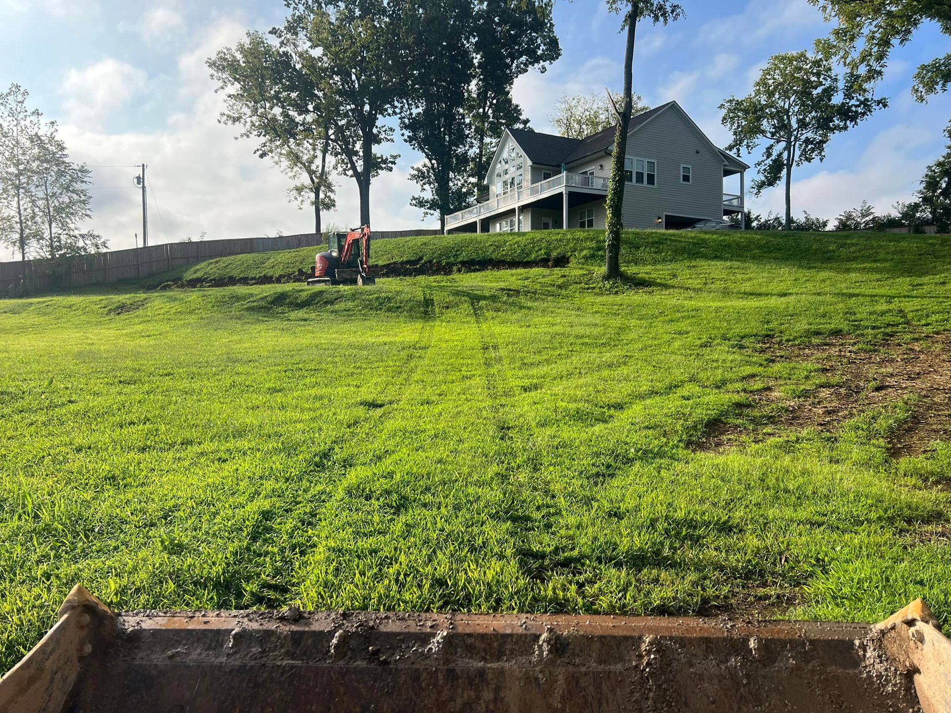 A front loader bucket filled with dirt on a grassy hill, a house sits on the hill's crest.