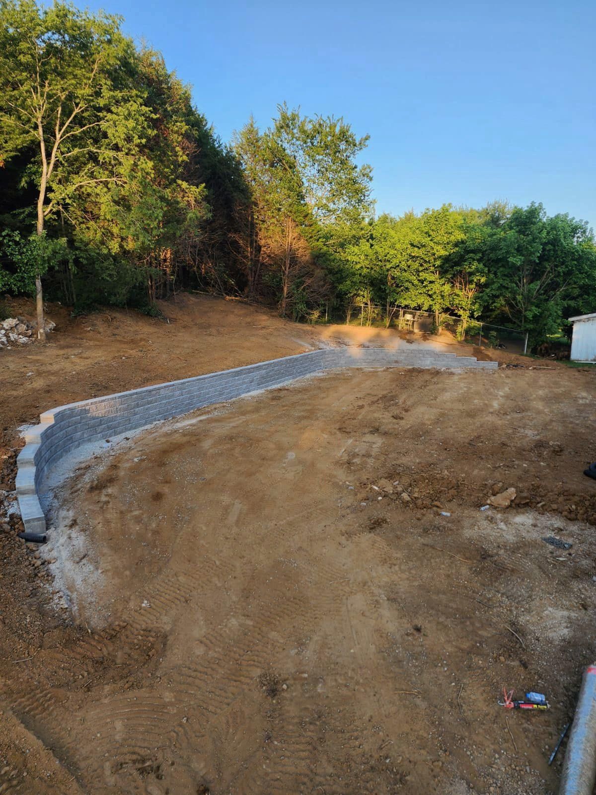 Construction site with a retaining wall made of gray blocks and gravel; dirt and trees surround.