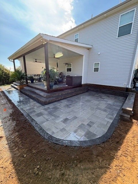 Newly constructed patio with gray pavers and a curved border next to a house with a covered porch.