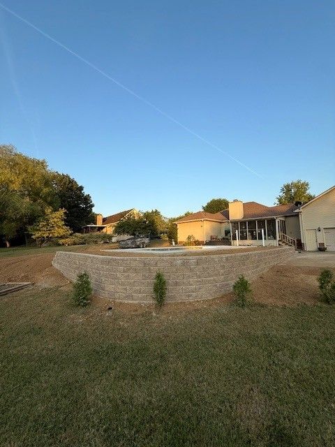 A backyard with a retaining wall, lawn, trees, and a house under a blue sky.