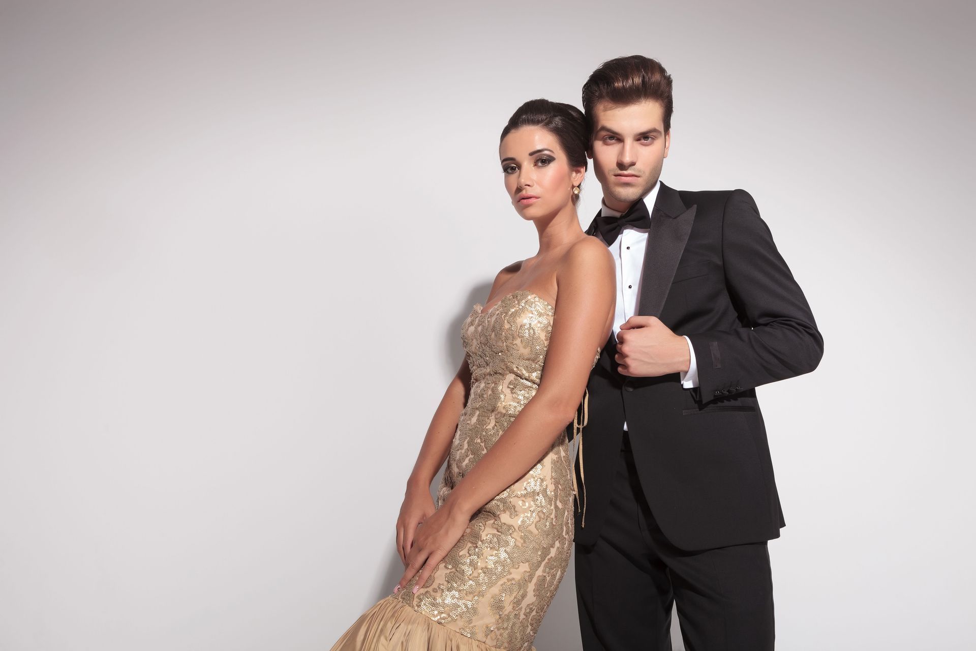 Woman in gold gown and man in tuxedo pose formally against a light gray backdrop.