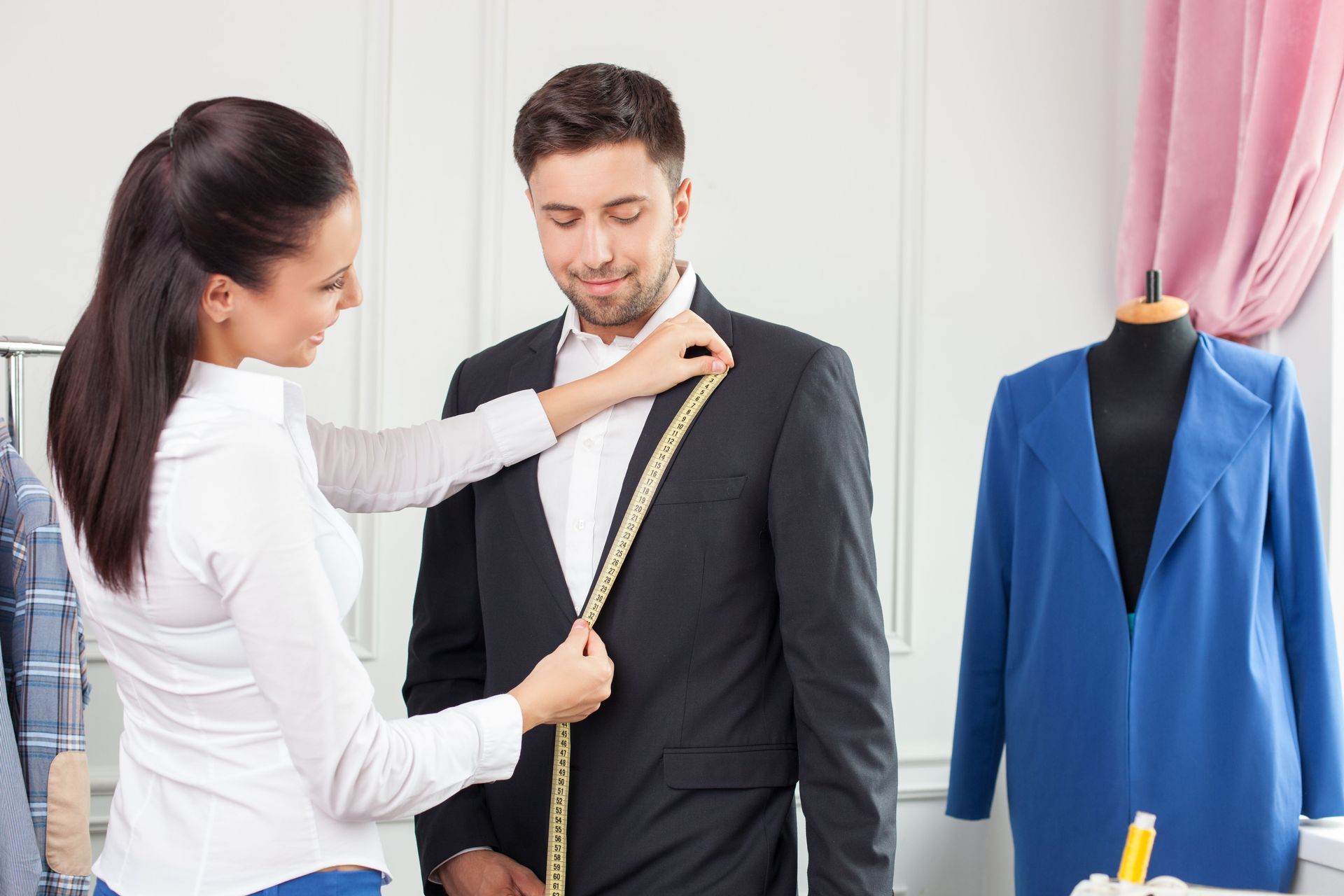 Woman tailor measuring a mans suit jacket with a measuring tape in a workshop.