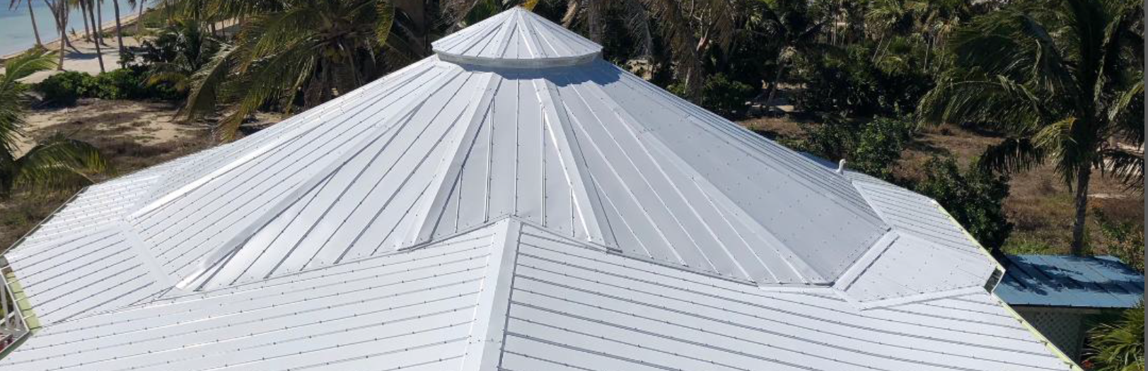 Aerial view of a white metal roof, with a center cupola, surrounded by trees and a hint of a beach.