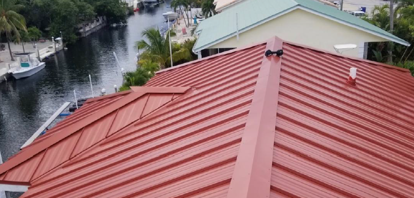Red metal roof of a house overlooking a waterway with boats. Trees and other houses in the background.