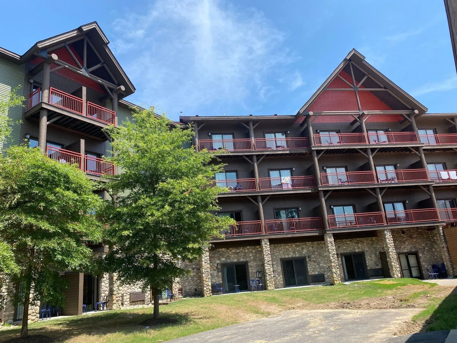 Multi-story lodge-style building with balconies, brown trim, stone base, and green trees against a blue sky.