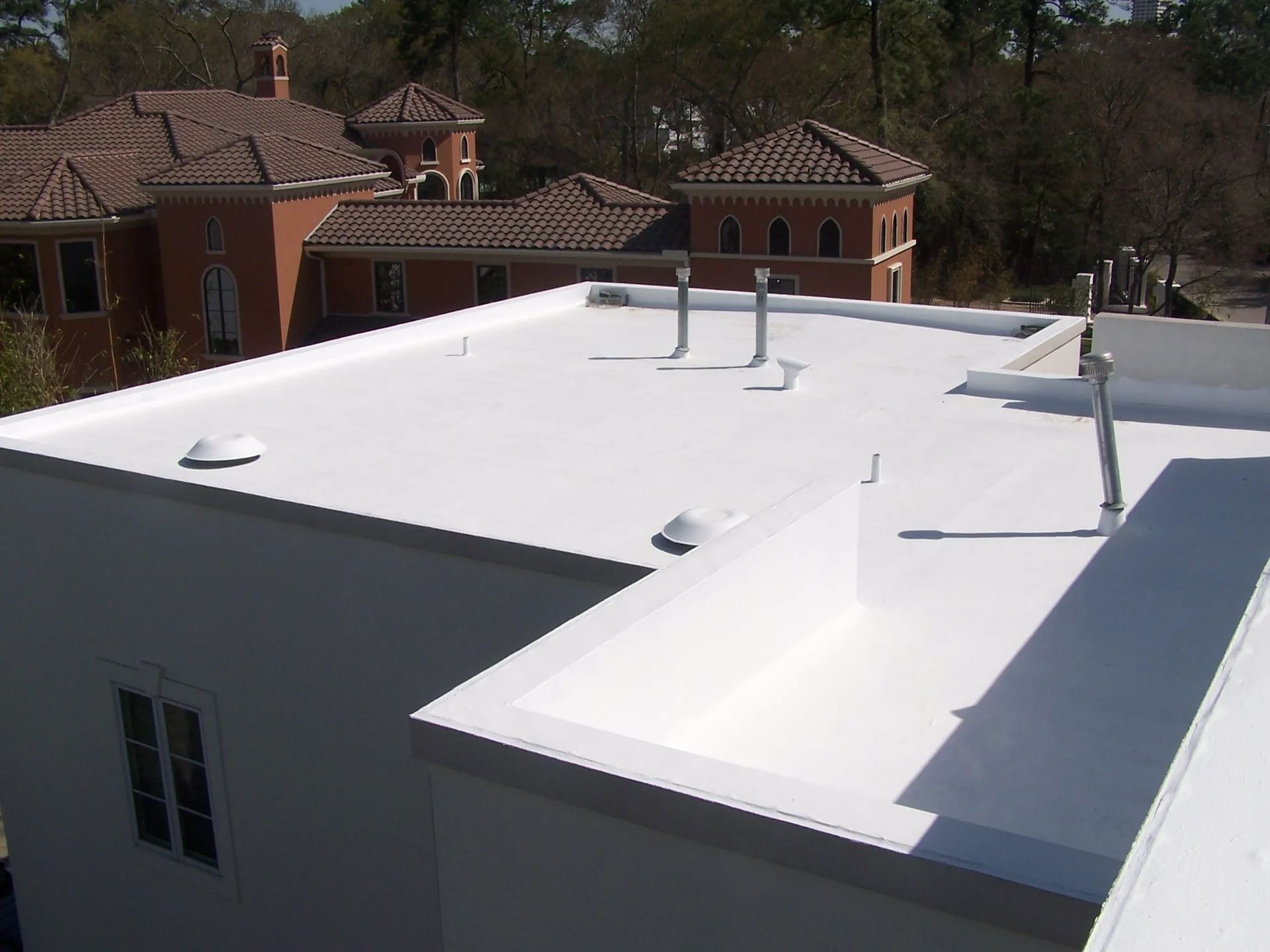 White flat roof on a building with multiple vents and a building with a red tile roof in the background.