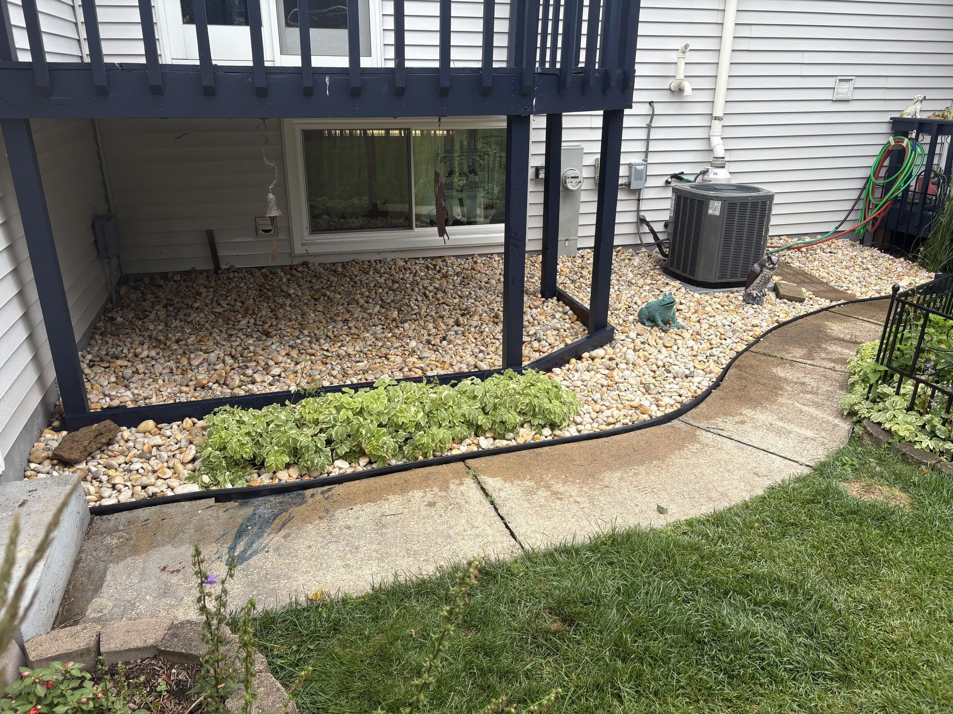 Concrete walkway alongside a rock garden under a deck, with an air conditioner unit and green grass.