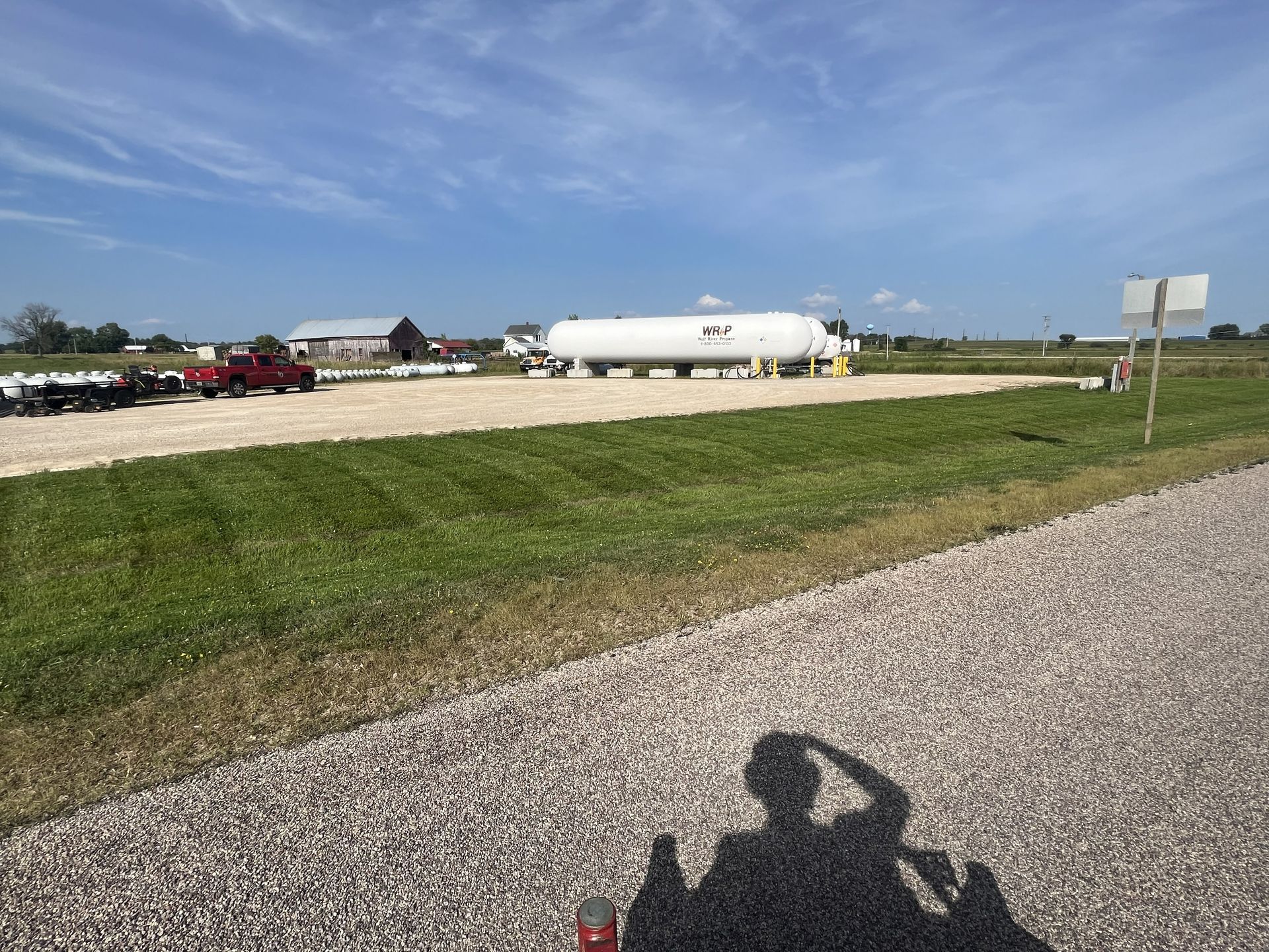 A large white tank in a grassy field with a gravel road. A shadow of a person is in the foreground.