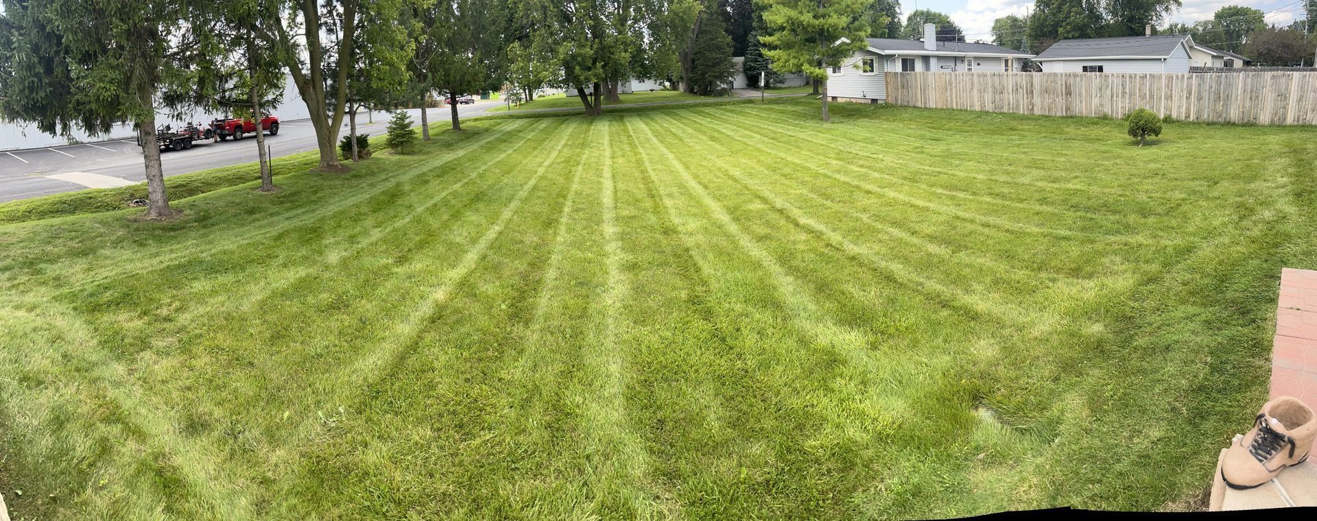 Lawn mowed with distinct stripes, trees and a fence in the background, a pair of sandals on the lower right corner.