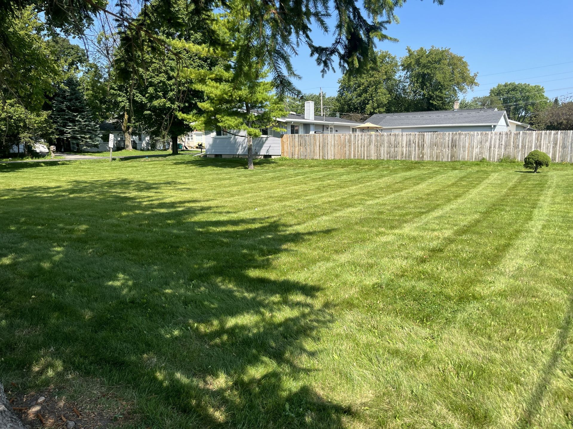Green grassy lot with freshly cut stripes; trees and houses in the background under a blue sky.