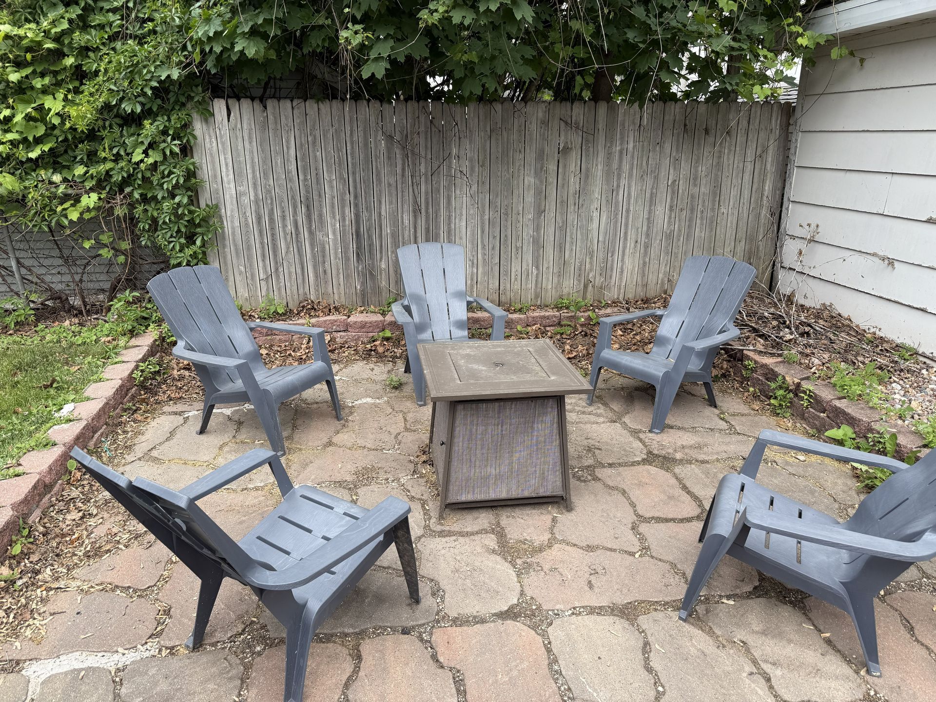 Five gray Adirondack chairs circle a small, weathered wooden table on a stone patio, enclosed by a wooden fence.