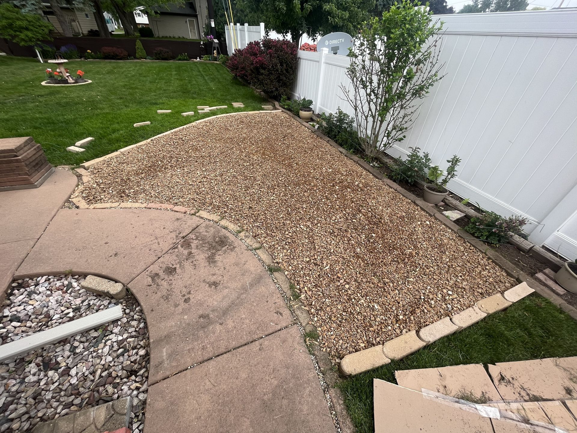 Gravel bed bordered by bricks next to a concrete patio and white fence, with green grass and shrubs.