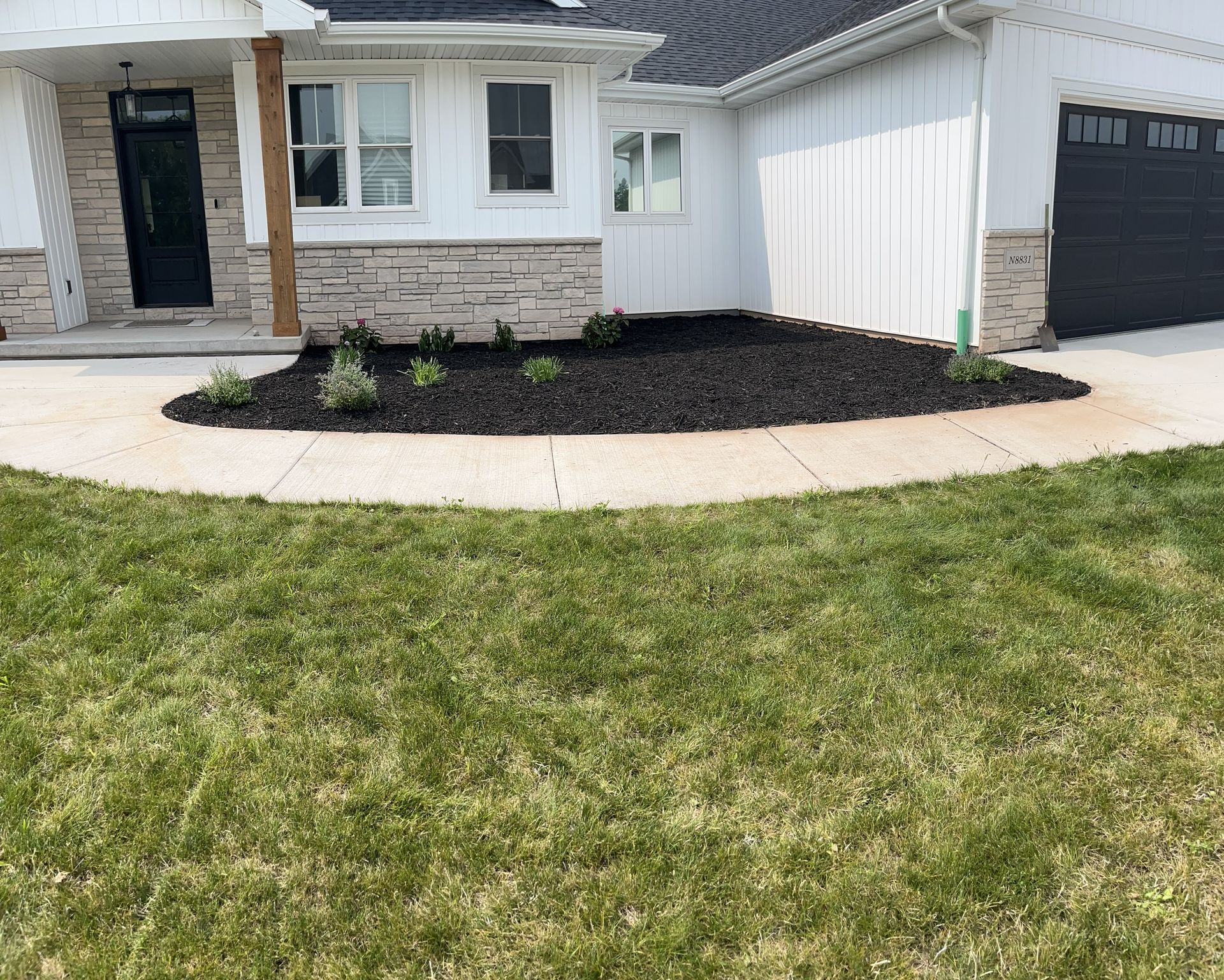 A front yard with a curved concrete walkway, flowerbed with dark mulch, and a white house.