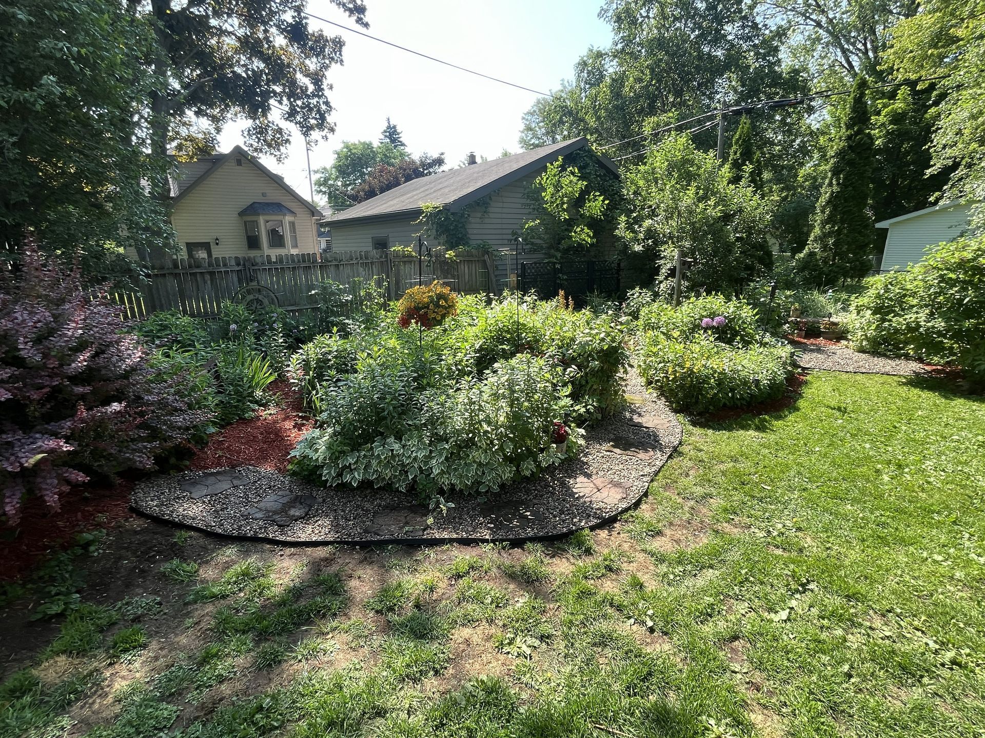 Backyard garden with green grass, various bushes, red mulch, and a house in the background.