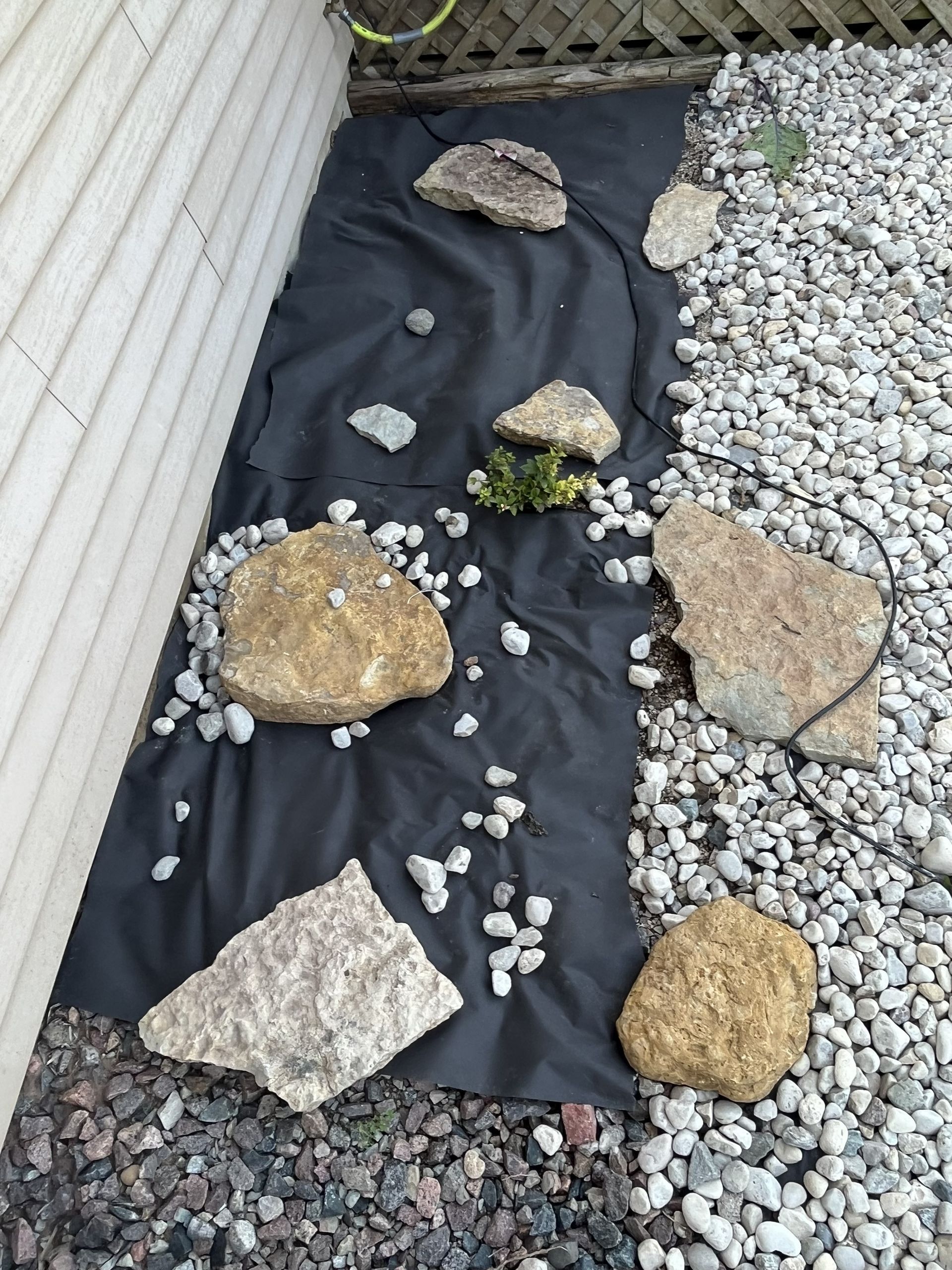 Black landscape fabric with rocks and a small green plant, next to gravel and a house wall.