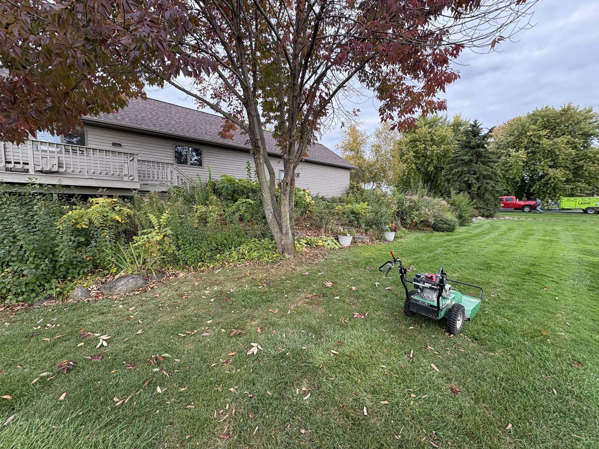 A lawnmower on grass next to a house with a tree and foliage. Cloudy sky.