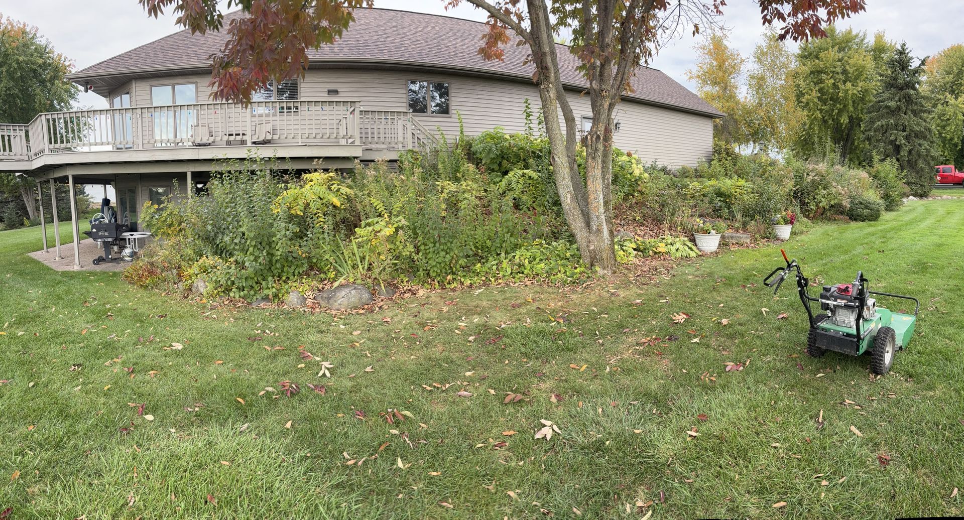 Lawnmower on a grassy hill next to a house with a deck and landscaping. Someone stands on the deck.