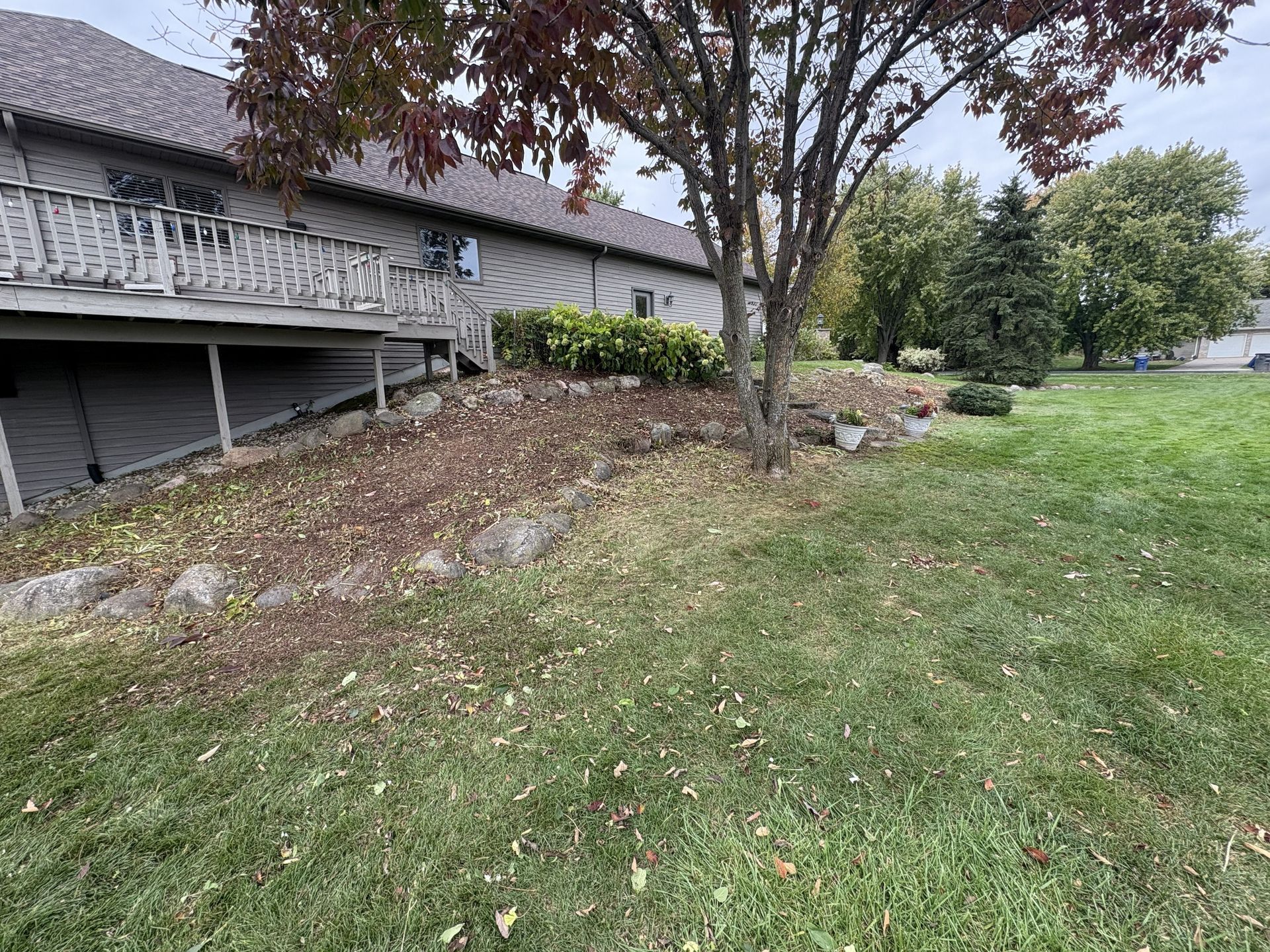 A house with a wooden deck next to a tree and a grassy yard on a cloudy day.