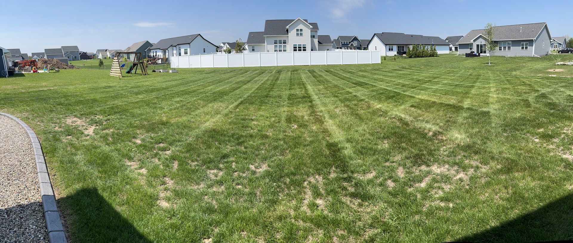 A wide shot of a green lawn in a suburban neighborhood, with a white fence and houses in the background.