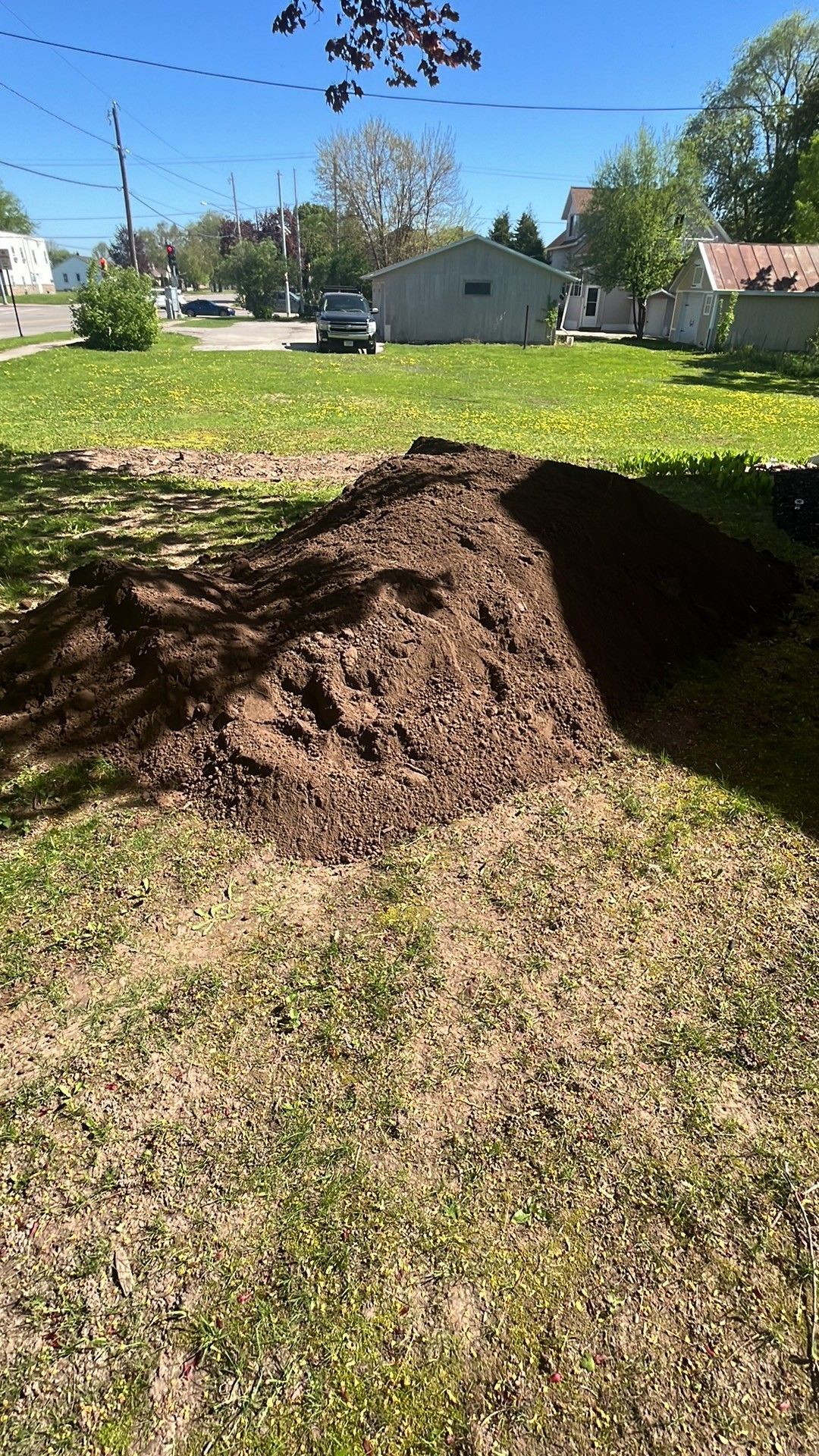 Large pile of brown mulch on a grassy lawn with houses and a vehicle in the background.