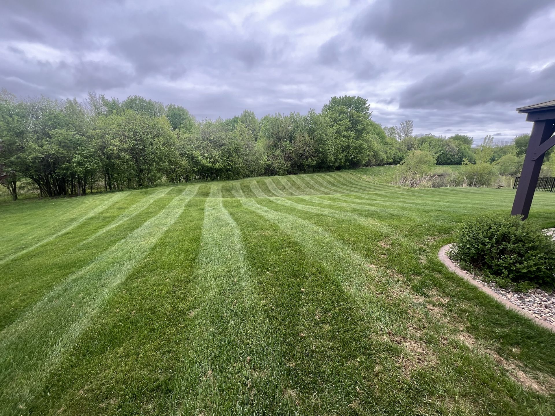 Lawn with alternating stripes, under a cloudy sky. Trees and a patio are visible in the background.