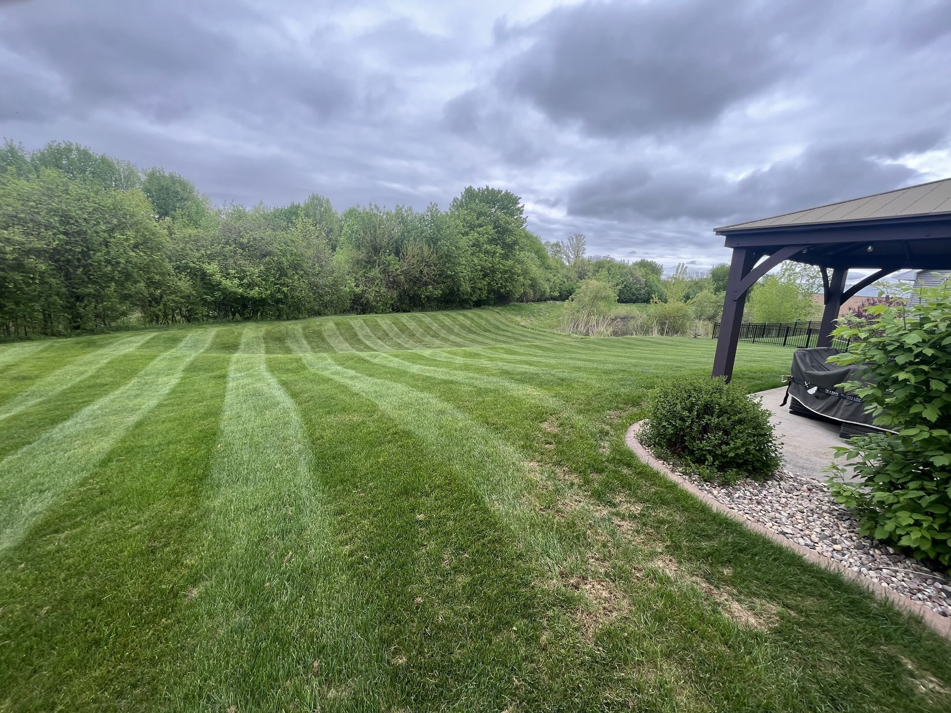 Green lawn mowed in stripes under a cloudy sky, with a gazebo and trees in the background.