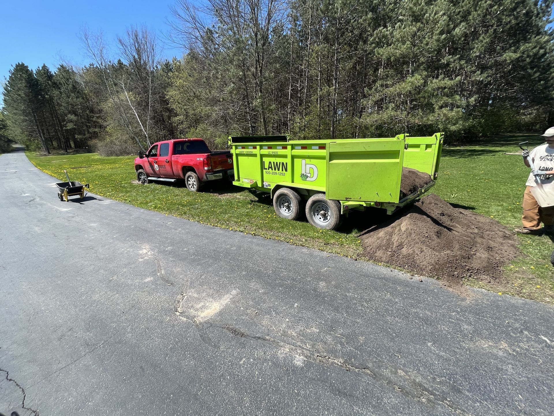 Red truck towing a bright green dump trailer, dumping soil onto the roadside. A dog is nearby.
