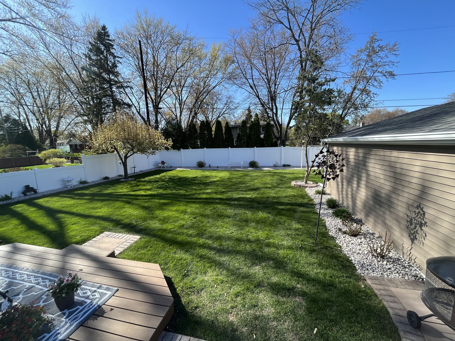 Backyard with green grass, white fence, trees, and a wooden deck. Sunny day.