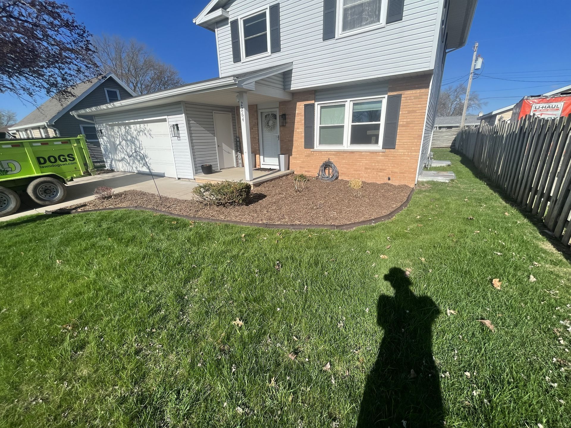 Two-story house with green lawn and mulch beds.  A shadow of a person is cast on the grass.