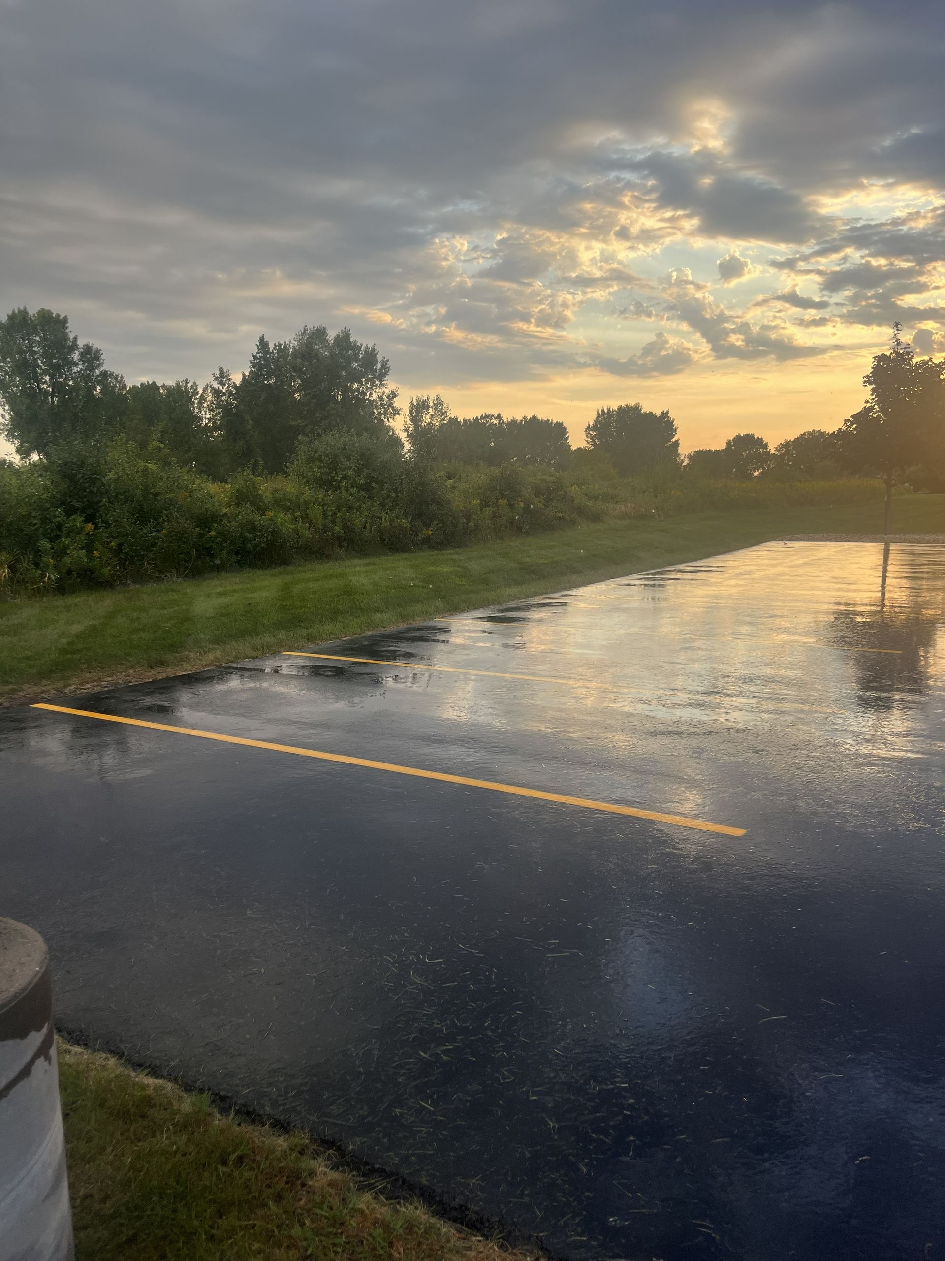 Wet asphalt road reflecting sunset, green grass, and trees under a cloudy sky.