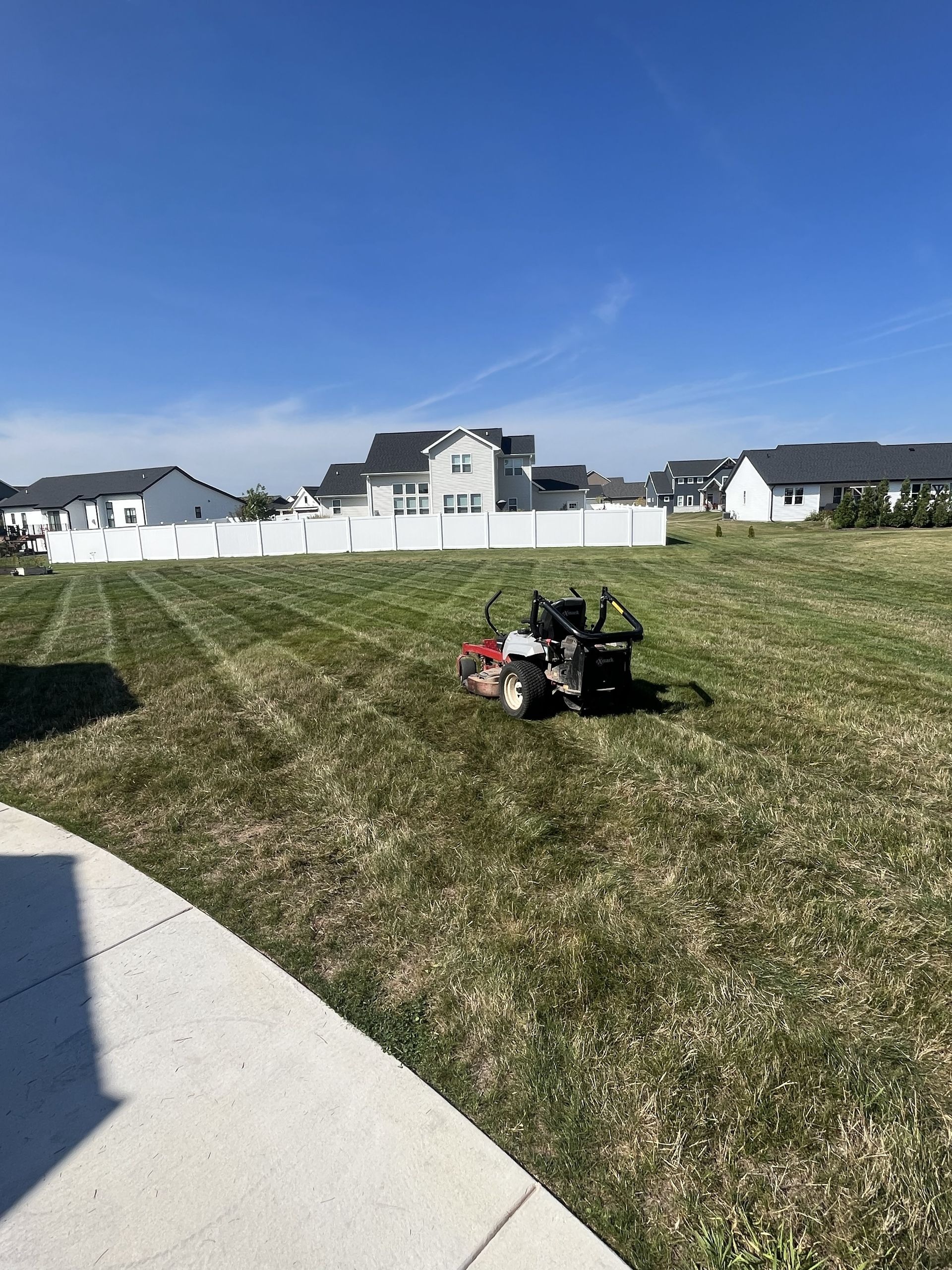 Lawn mower cutting grass in a backyard on a sunny day. Houses and a white fence are in the background.
