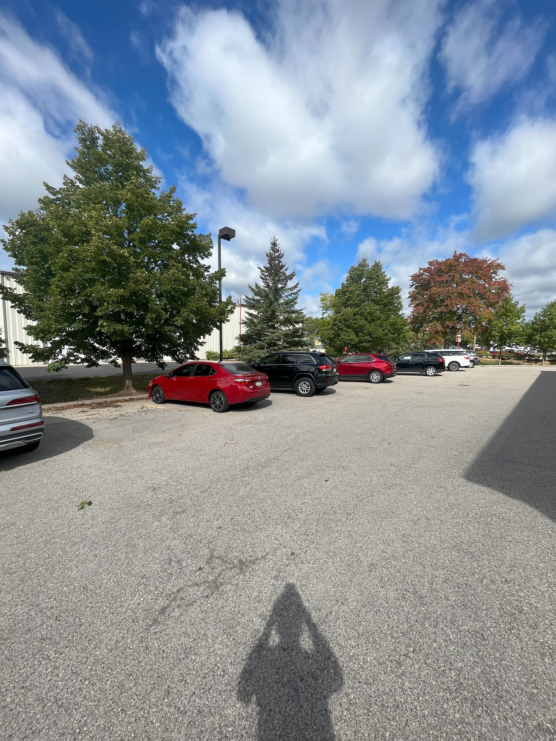 Cars parked in a gravel lot under a partly cloudy sky, shadowed figure in foreground, trees in background.