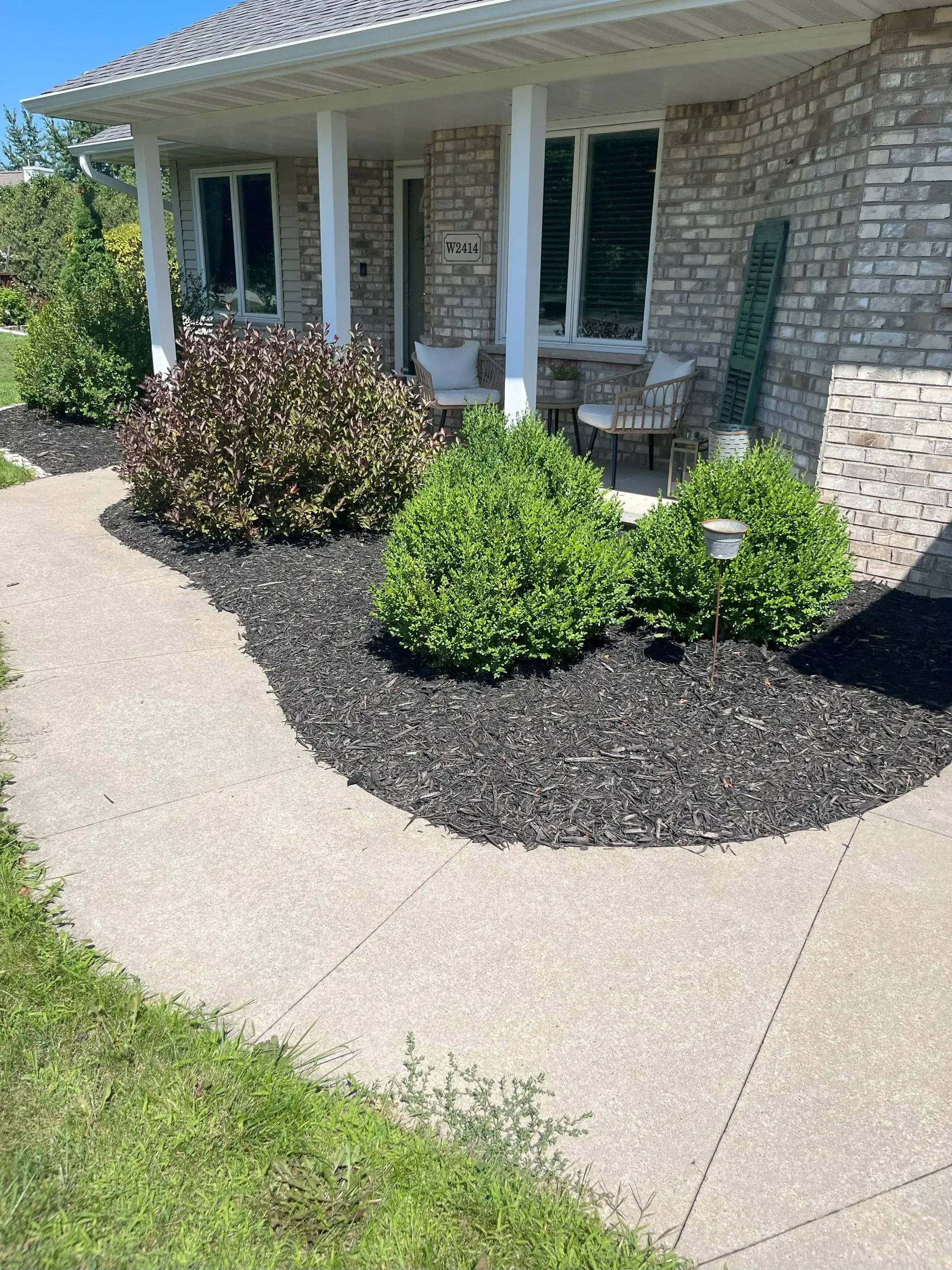 A paved walkway leads to a brick house with bushes and black mulch.