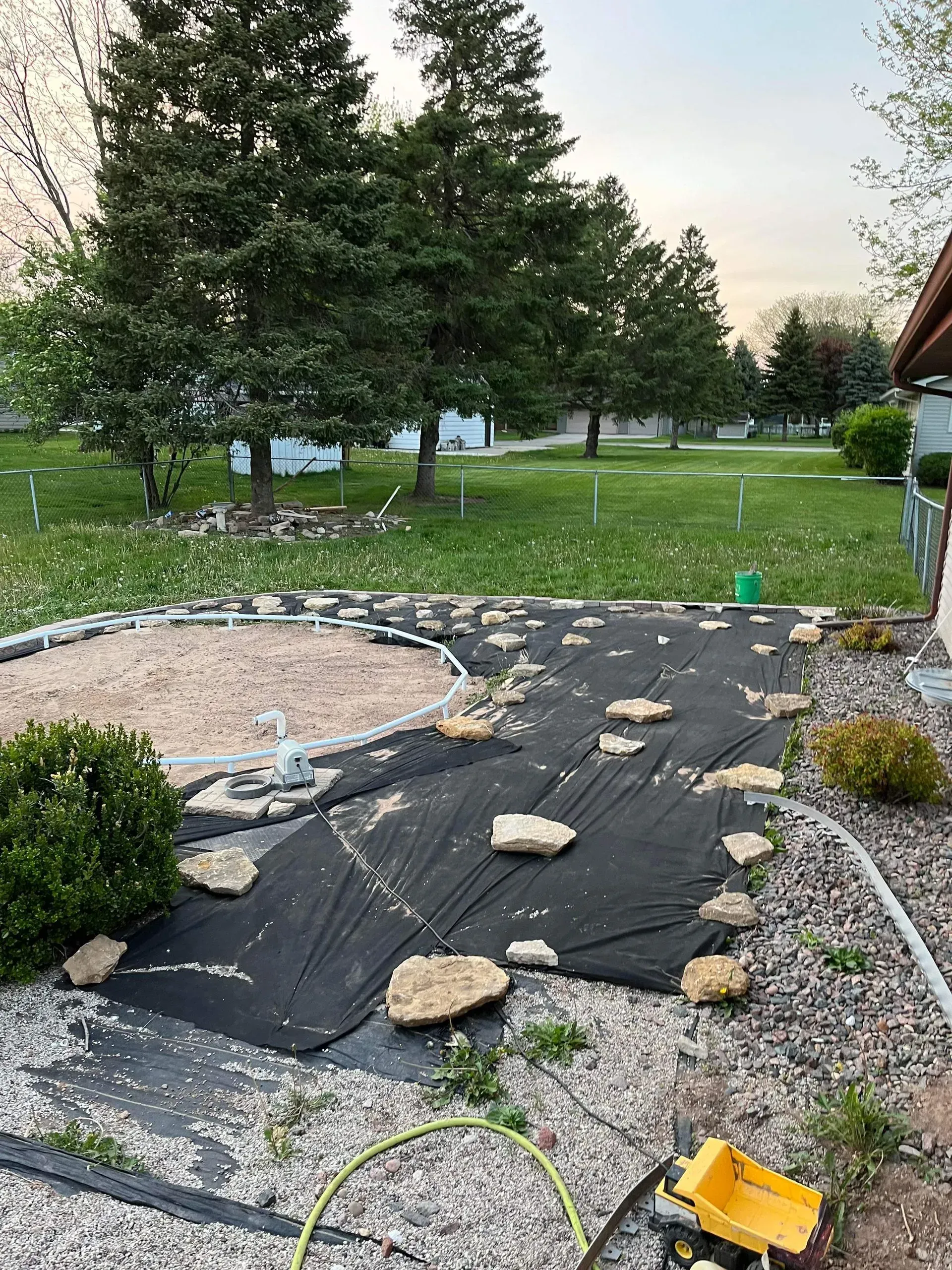 Black landscape fabric held down with stones covers a garden bed. Gravel and a sandbox border the area.