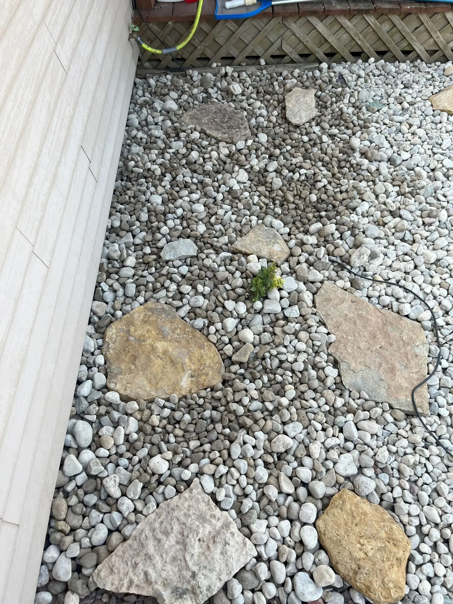 Gravel ground cover with large stones near a building's white siding. A green hose is visible above.
