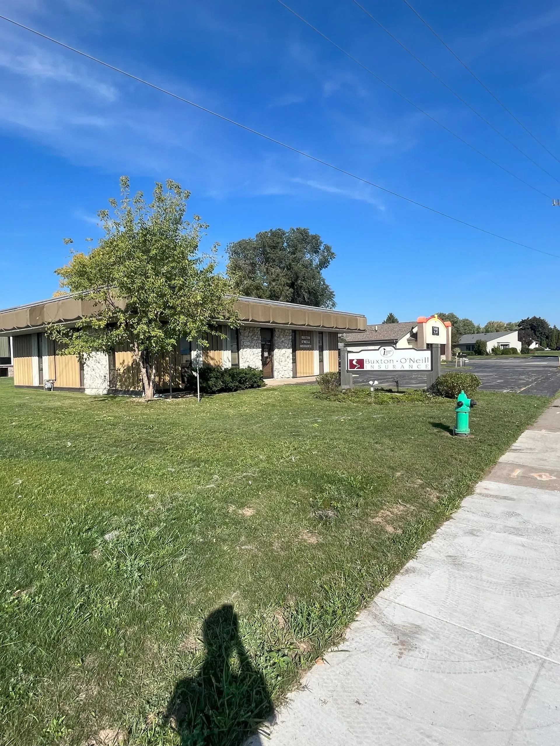 Single-story building with tan and white facade, surrounded by grass and a sidewalk. Tree in front. Blue sky.